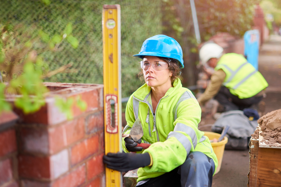 A construction worker wearing a green high vis jacket, blue hard hat, measuring how level a wall is