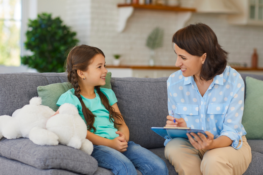 A professional conversing with a child as they write on a clipboard, whilst sat on a sofa