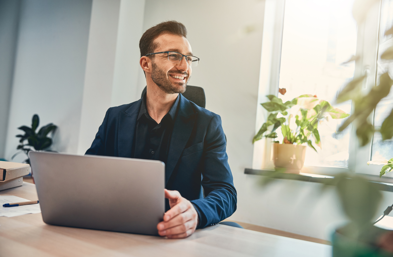 A professional wearing glasses and a navy blue blazer, holding a laptop whilst sat at a desk