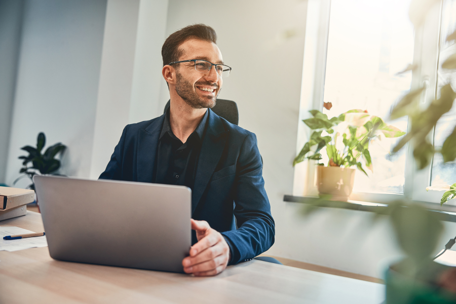A professional wearing glasses and a navy blue blazer, holding a laptop whilst sat at a desk