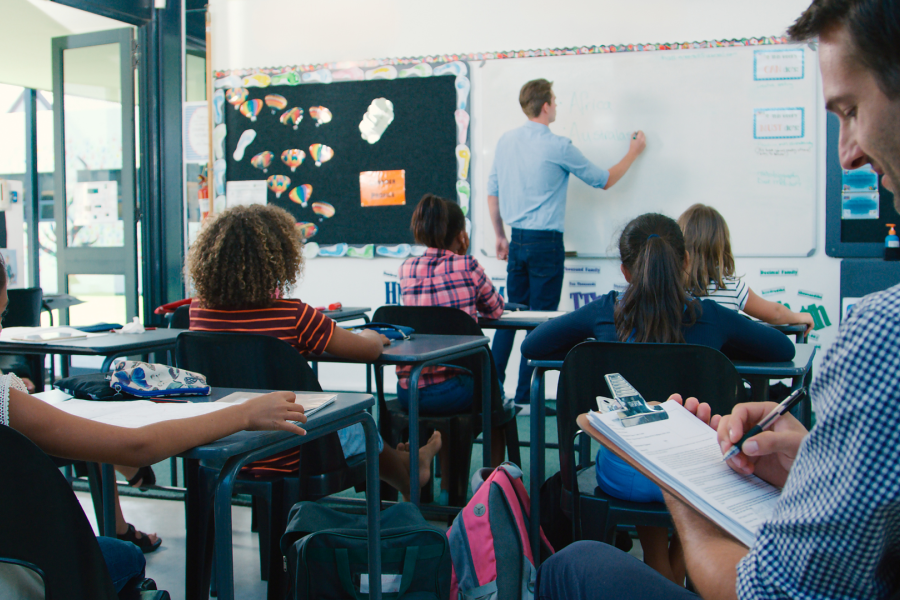 An adult sat at the back of a classroom and a teacher at the front writing on a whiteboard with a group of young children watching