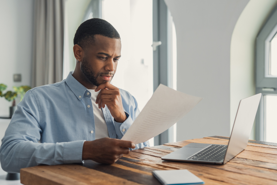 An adult sat down looking at a piece of paper with a laptop in front of them