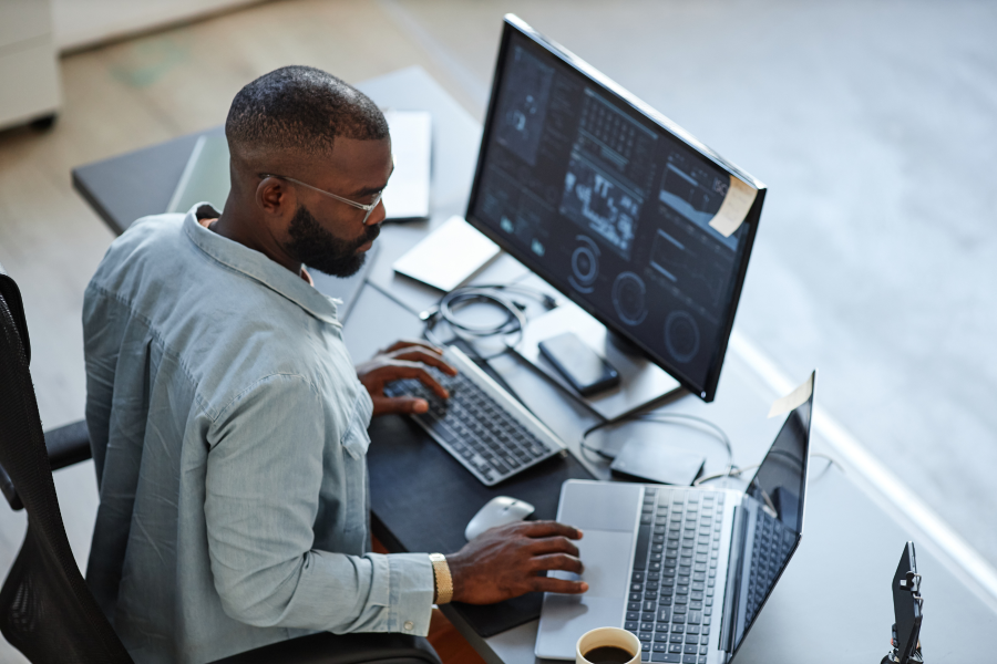 A professional sat at a desk using a desktop screen and laptop