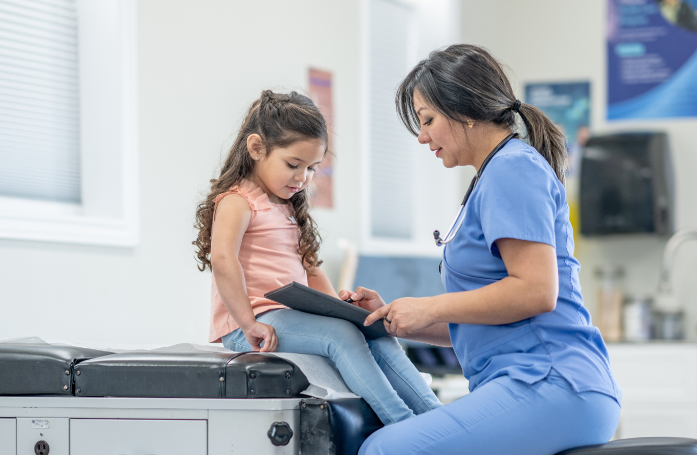 A healthcare professional wearing blue scrubs sitting with a young child, looking at a notebook