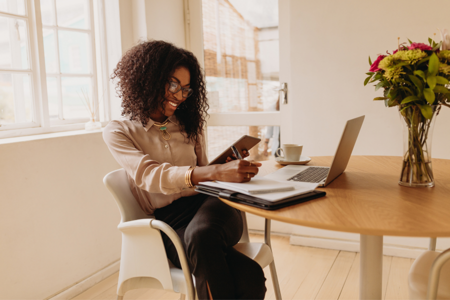 A smart professional sits with a laptop, writing on a notepad and looking at a digital device