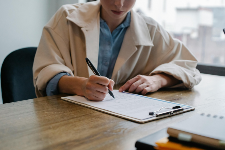 The torso of a person sat at a desk writing on paper attached to a clipboard