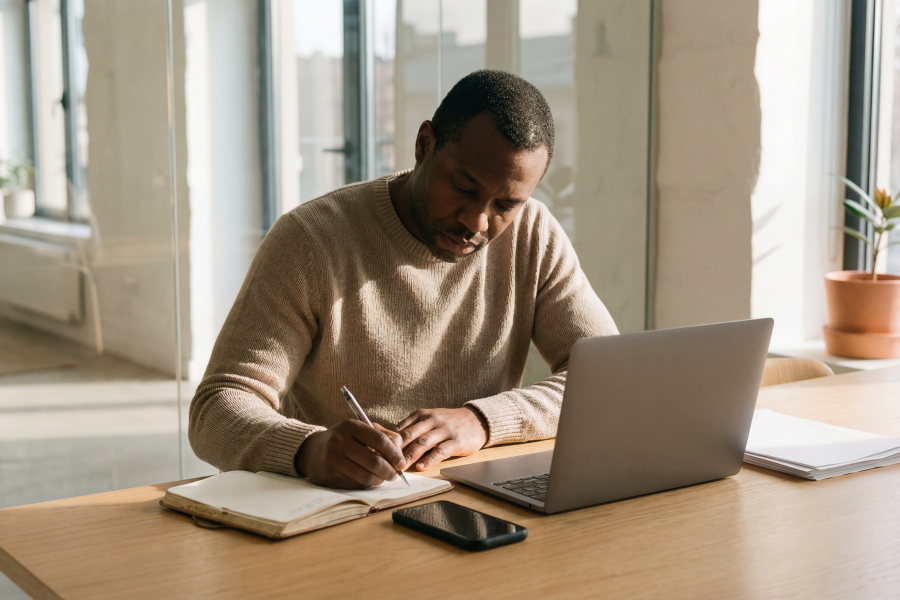 A professional wearing a beige jumper, writing into a notebook by an open laptop