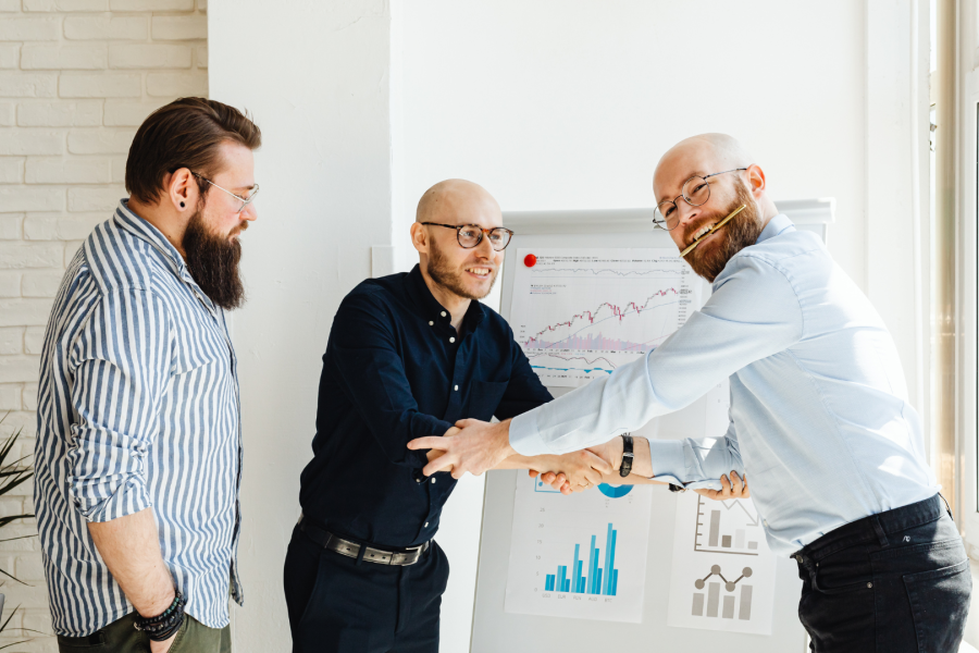 Three professionals in the office smiling, two are shaking hands and supporting eachother
