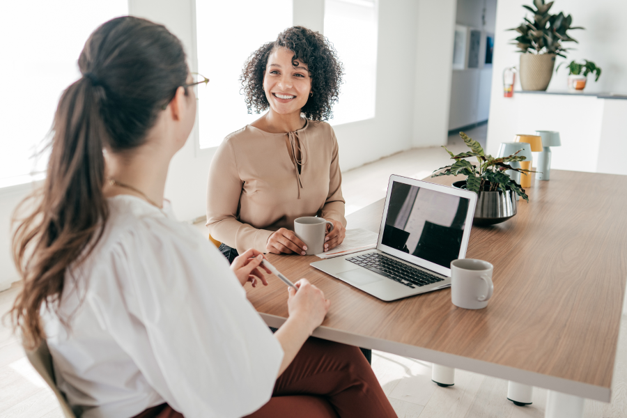 Two professionals sat at a desk talking and drinking out of mugs