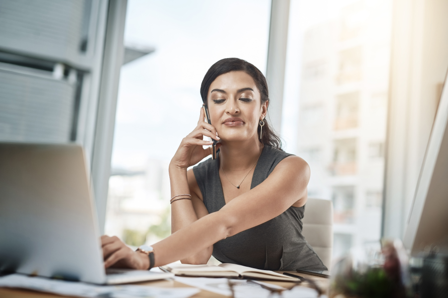 A professional using one hand to type on a laptop and the other hand to hold a mobile phone to their ear