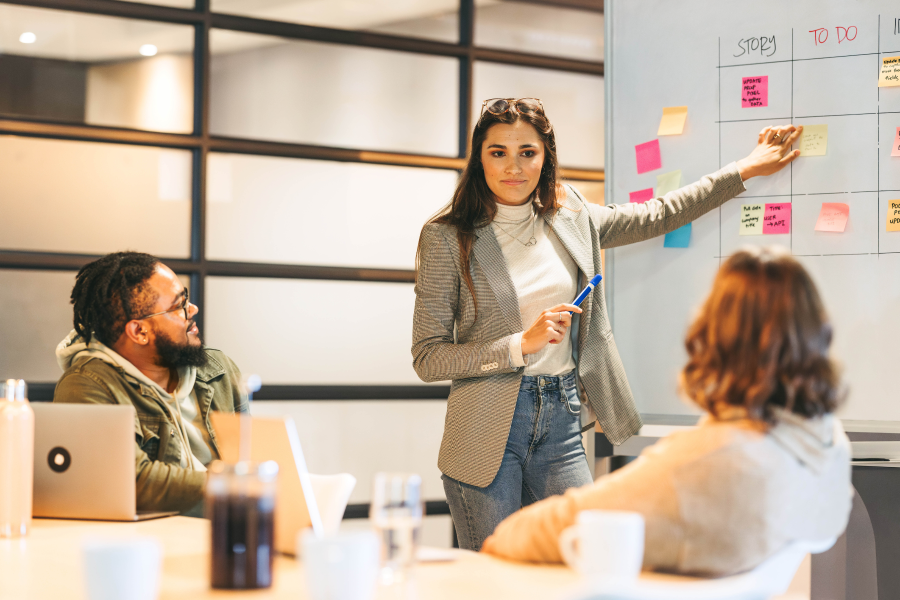 Two professionals sat whilst another person points at a whiteboard with notes stuck to it