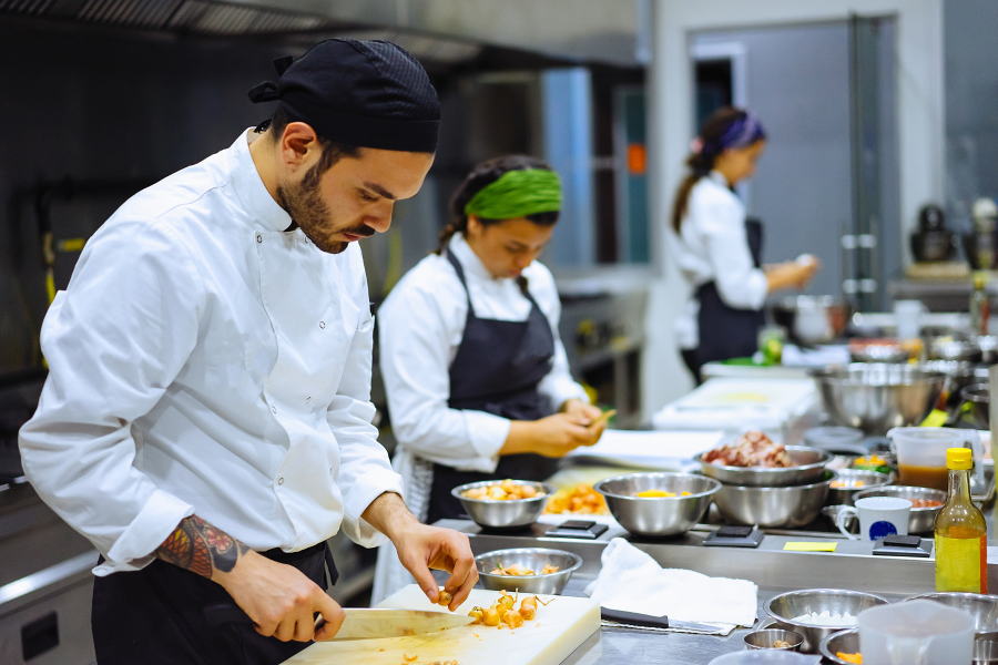 Three cooks stood in a kitchen preparing food whilst wearing chefs uniform