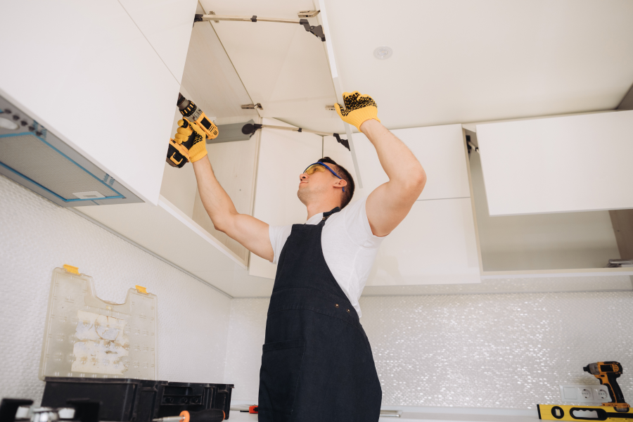 A tradesperson wearing black overalls and a white t-shirt, drilling a cupboard together
