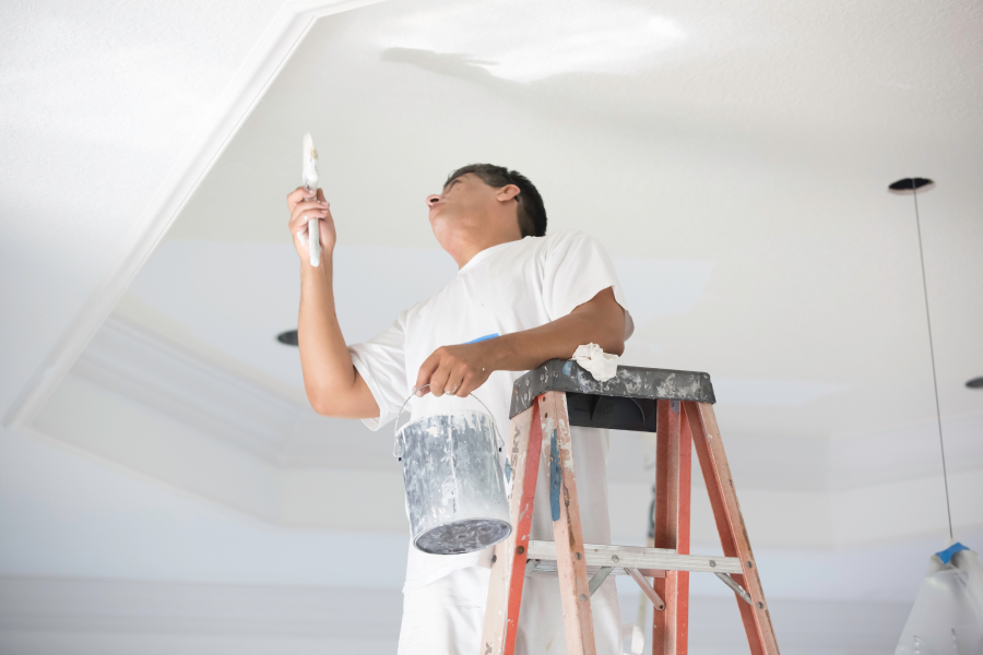 A tradesperson stood on a red ladder, painting a ceiling in white paint