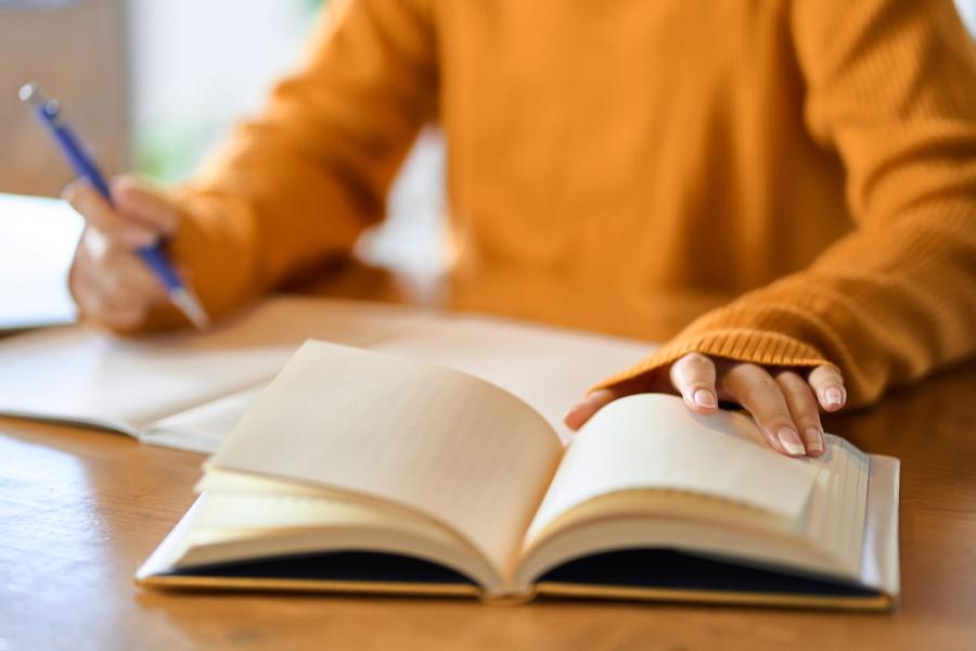 An individual writing on a piece of paper and holding a book