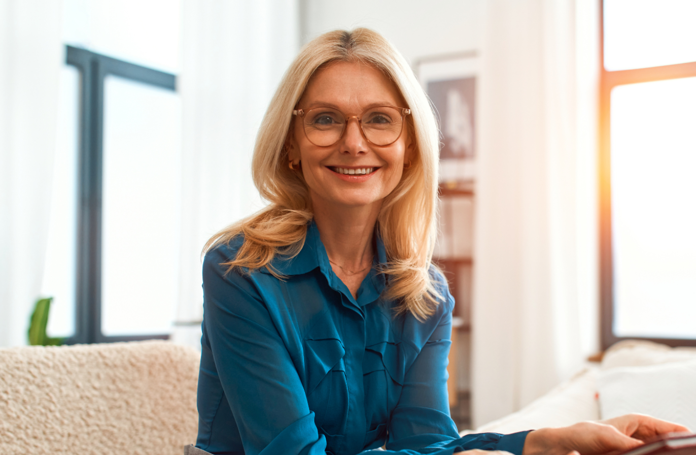A professional wearing a blue shirt, grey trousers and glasses, sitting down smiling