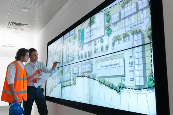 A man in a suit and a man wearing high vis looking at town plans on a screen